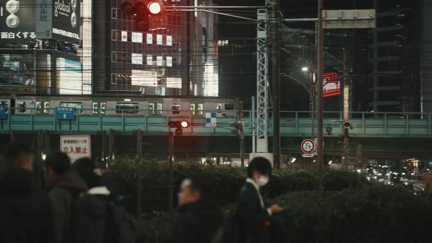 Night urban scene with people crossing the street moving cars and illuminated buildings in the background and train passing on an overpass