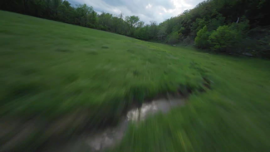 An aerial view of a green field with a small stream that flows into a larger river in the middle of a forest