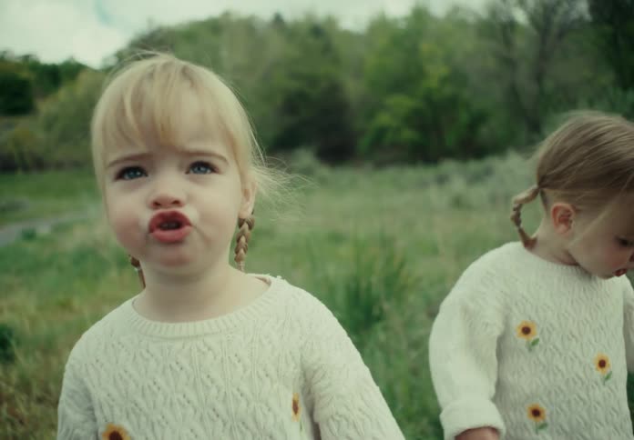 Two little blonde girls in white coats are walking on a lawn and looking straight ahead one of them is pouting