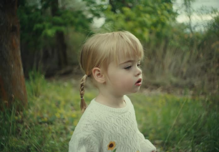 A blonde toddler stands on grass in the woods staring and thinking