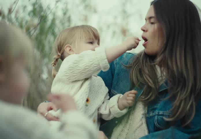 A toddler pokes her mother with a plant stem while her sister is nearby