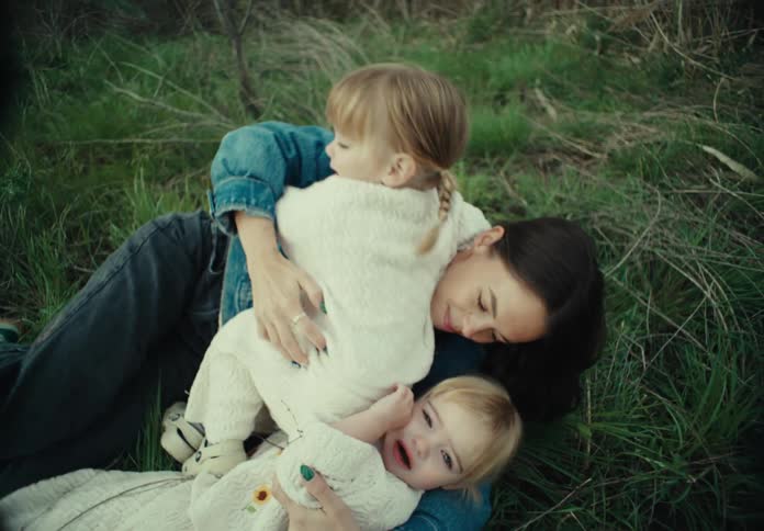 A mother lies down outside in the grass with her twin daughters