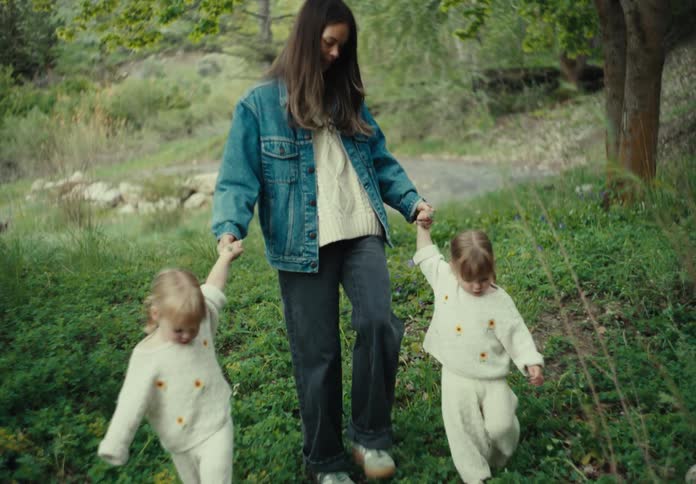 A mother walks her two toddler daughters over a grassy area near a tree