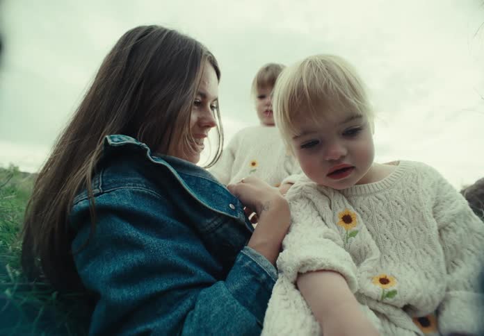 A little blonde child on the lap of an adult woman in jeans and she is talking to a little child and she is happy and there is another child behind them