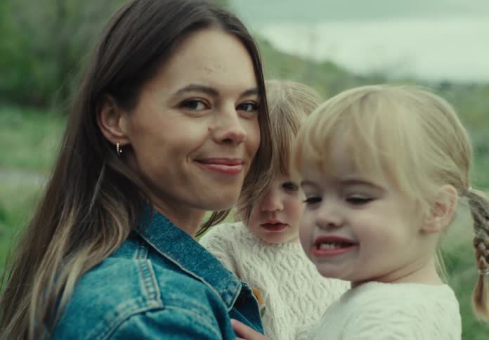 Mother smiles while hugging her daughters in a field near a hill on an overcast day
