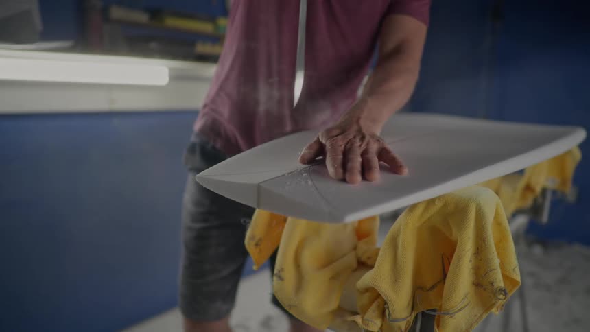 Man working in a workshop uses a blade to shape a flat white object into a surfboard