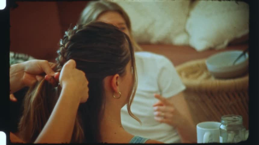 Pair of hands braiding the hair of a woman who is sitting talking to another woman next to her