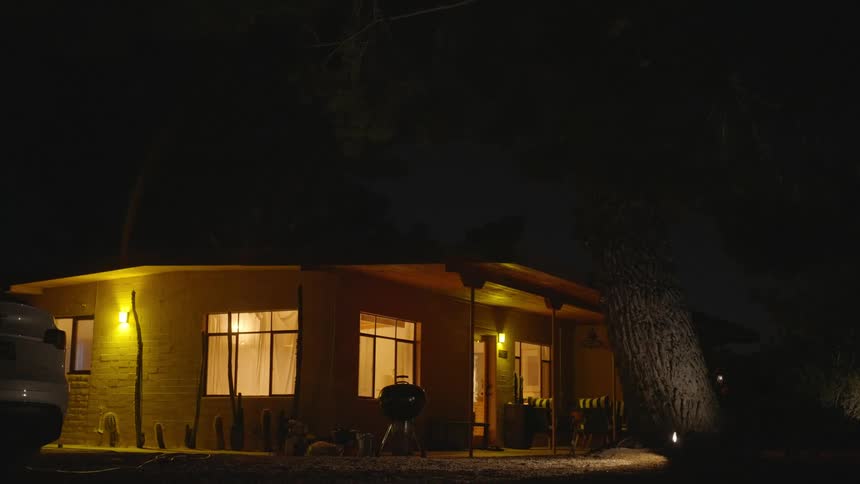 A nighttime scene of a singlestory house with illuminated windows and a porch light