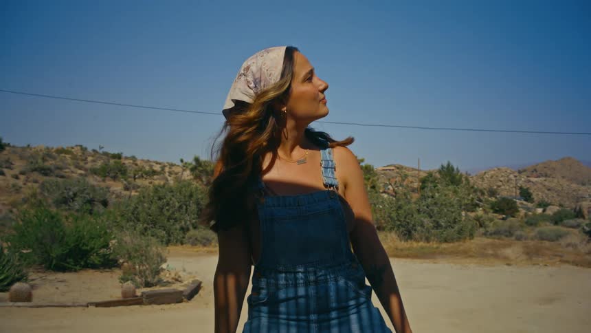 A young tan woman stand outdoors in a desert while smiling at someone