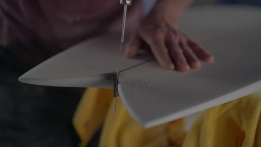 Closeup of a persons hands working with a thin pointed tool scraping the back of a surfboard