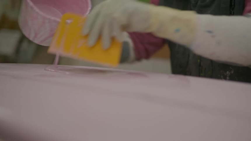 Hand with gloves and a spatula passing a pink liquid above a surfboard