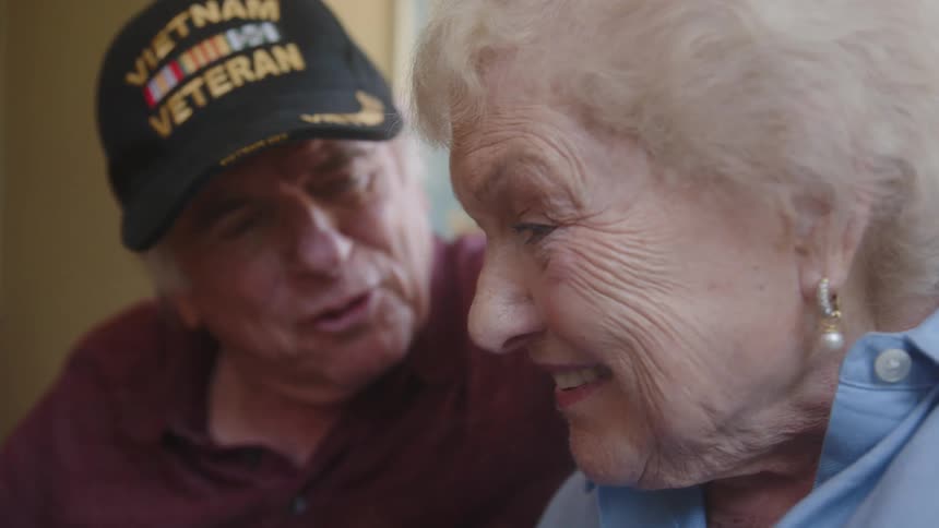 An elderly man and woman stand next to each other and are looking forward and laughing and talking