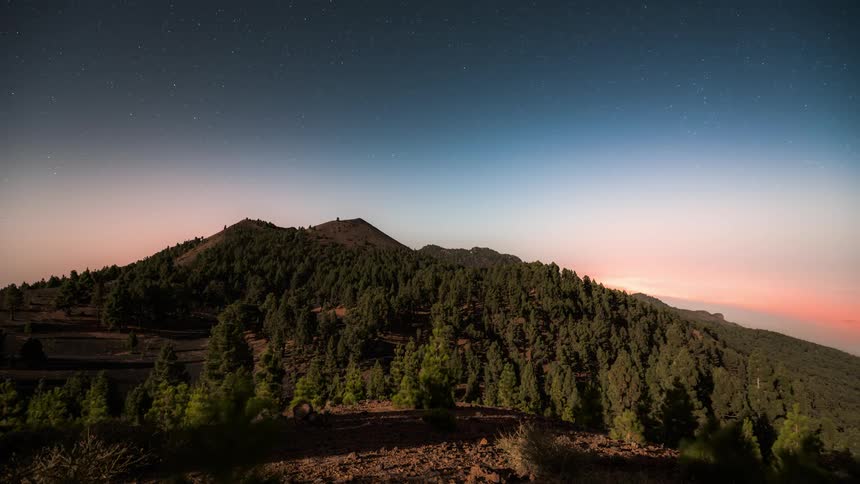 Day sky becomes night sky over a forested mountain as day and night pass in turn