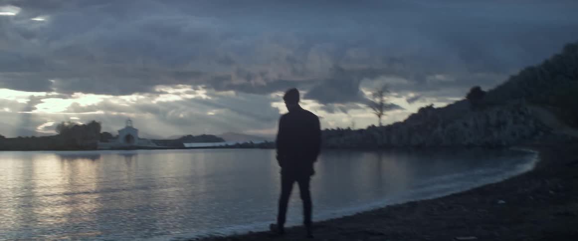 A man walking on the shoreline towards the water and then bending down to pick something up on a very cloudy day