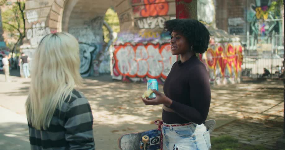 Two skater girls stand beneath an archway decorated with graffiti and talking