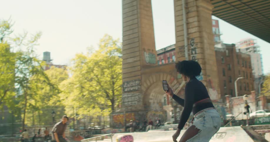 A black woman on a skateboard films another skater passing her by in a city park