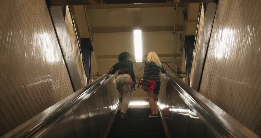 Two girls carrying skateboards riding up the escalator from a terminal
