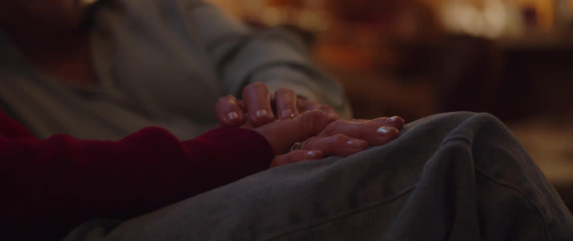 An elderly couple sitting on a couch at home holding hands while watching television