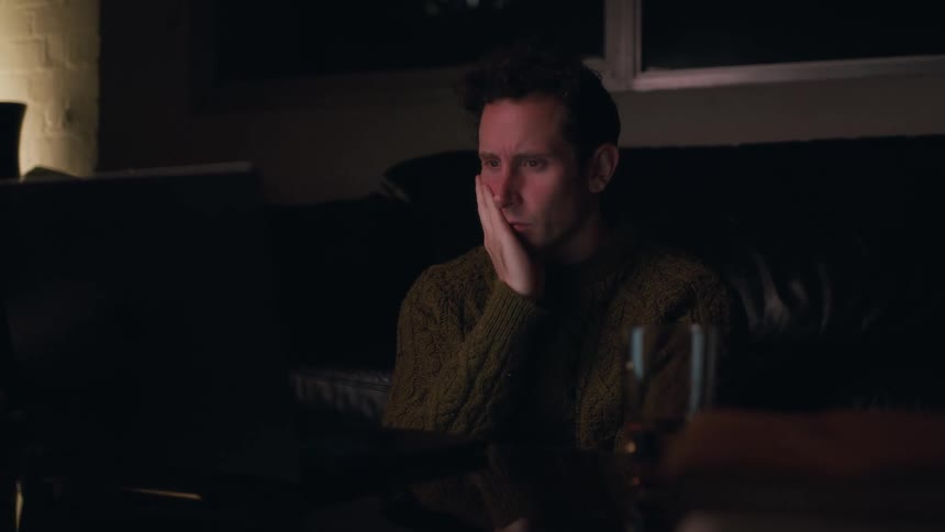 A man sitting in a dimly lit room tired and impatiently waiting in front of a laptop