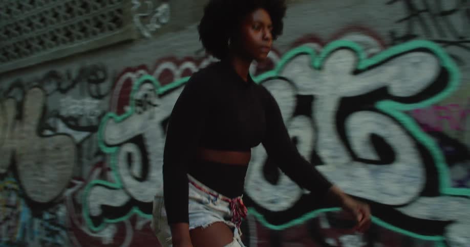 A young black skater girl riding her skateboard past a wall of graffiti in the city