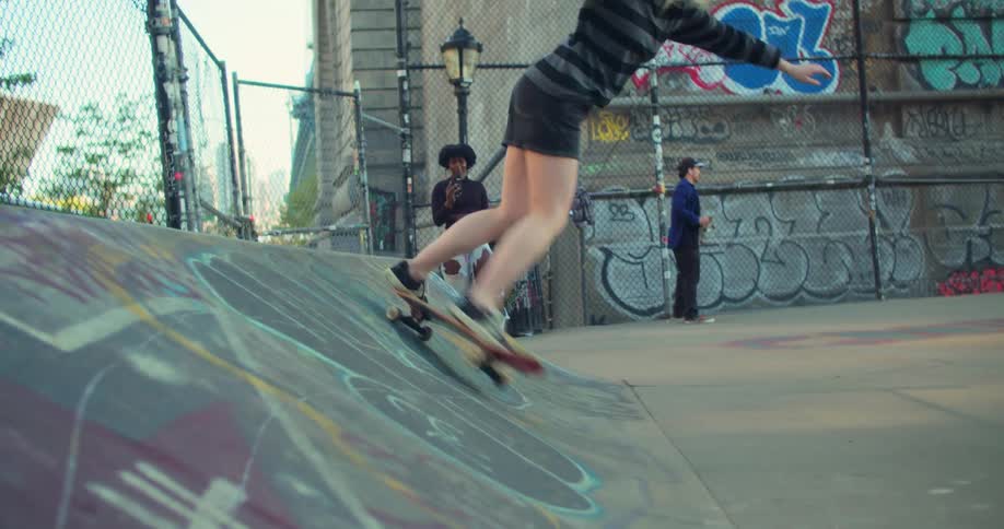 A girl rides a skateboard over a small ramp while a friend films with her phone