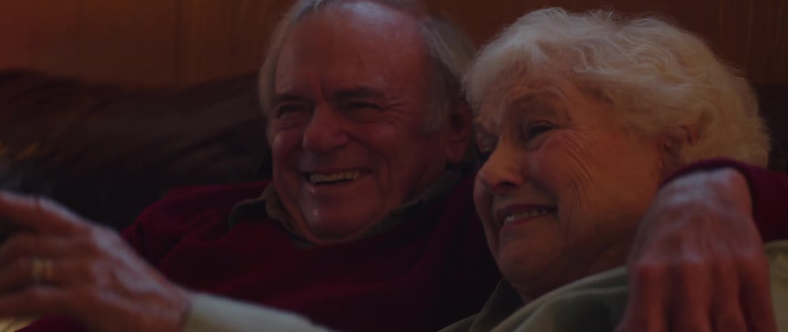 Elderly couple sitting in living room smiles while viewing television