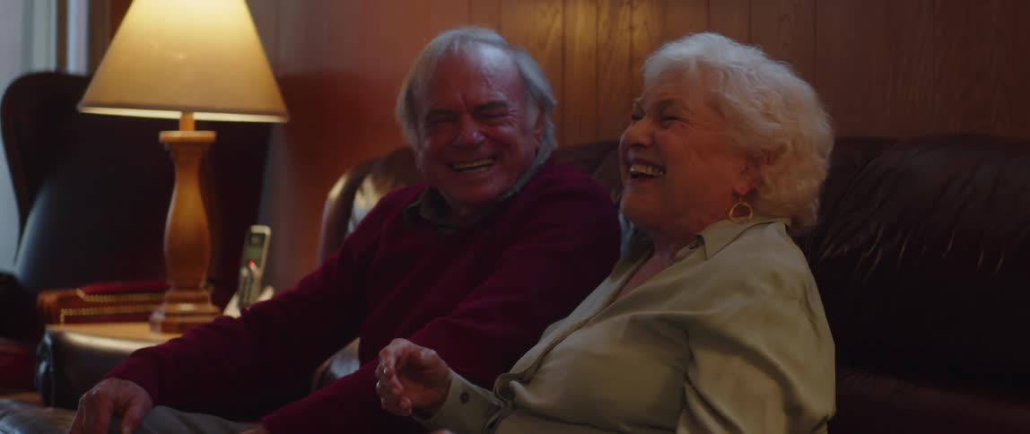 A happy elderly couple sitting on a couch at home laughing together while watching television