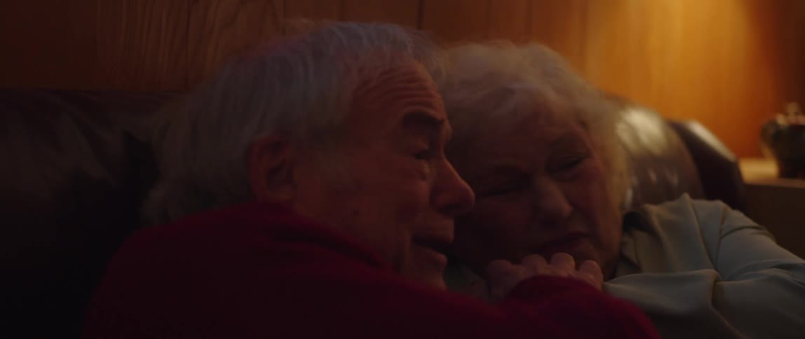 An elderly man and woman are sitting and watching something and the woman is scared and the man helps her