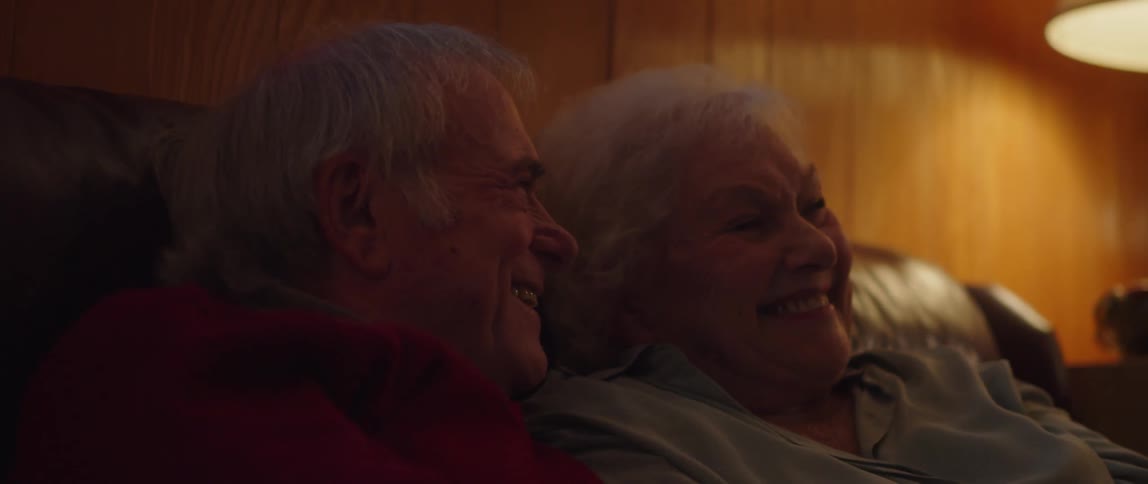 An elderly couple laughing and playing around with one another while sitting on a couch in a living room