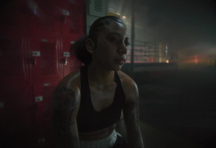 A young female boxer covered in sweat sits in front of red lockers under flashing strobing lights
