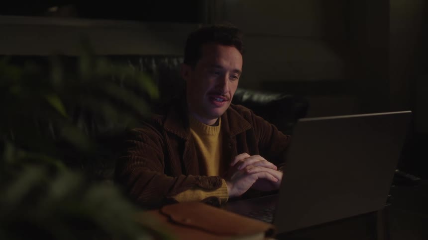 A young man is talking to his computer screen and he is sitting in a room