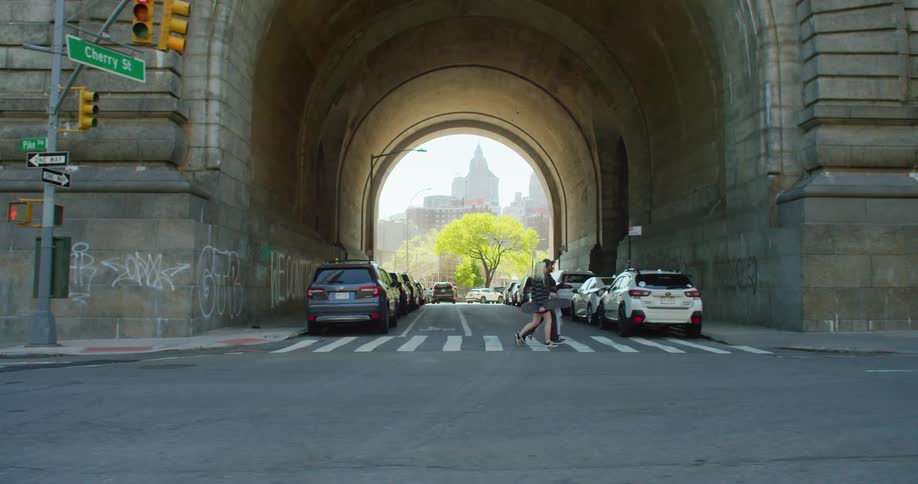 Street seen from inside a tunnel with parked cars on the right and a moving car and three friends crossing a street