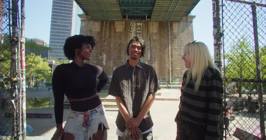 Three friends stand smiling holding skateboards in front of a skateboard park