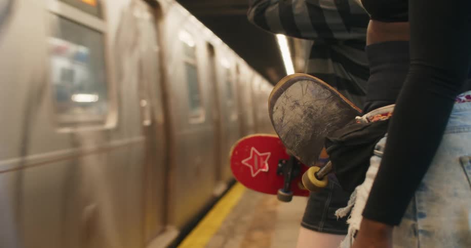 Two people each holding a skateboard while standing at a station with a train passing in front
