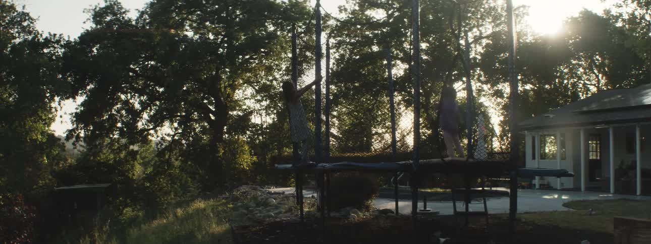 Two young female kids jump on a trampoline while dogs jump and bark nearby