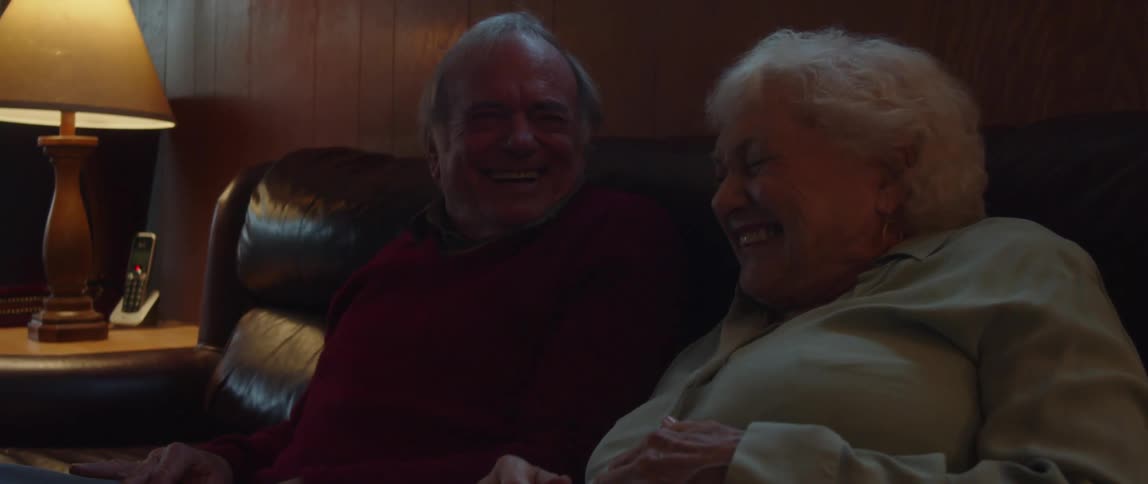 An elderly couple laugh with each other while sitting on a couch