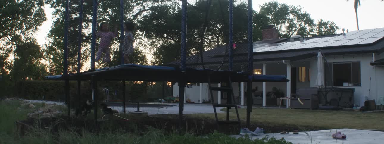 Two girls jump on a trampoline in a backyard as two puppies chase them around underneath