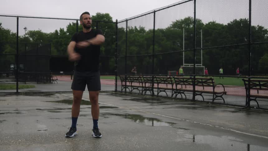A man stands on a tennis court and stretches his arms on a cloudy summer day