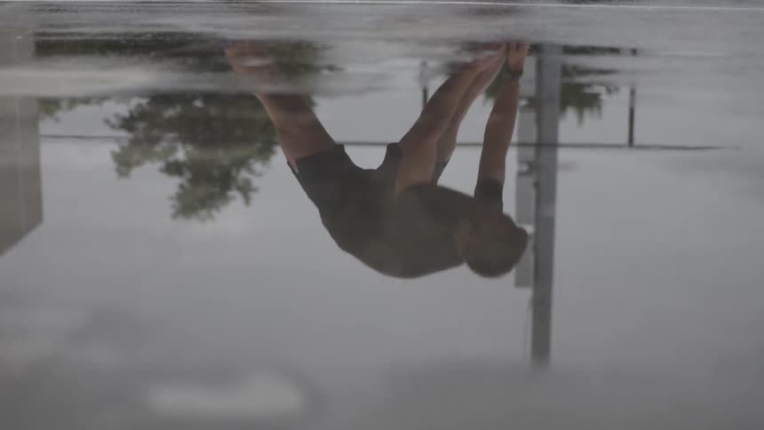 A young bearded man's reflection while performing warm up exercises on a wet court under an overcast sky