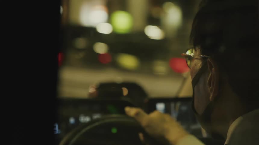 A manwearing glasses drives a car on a city street at night