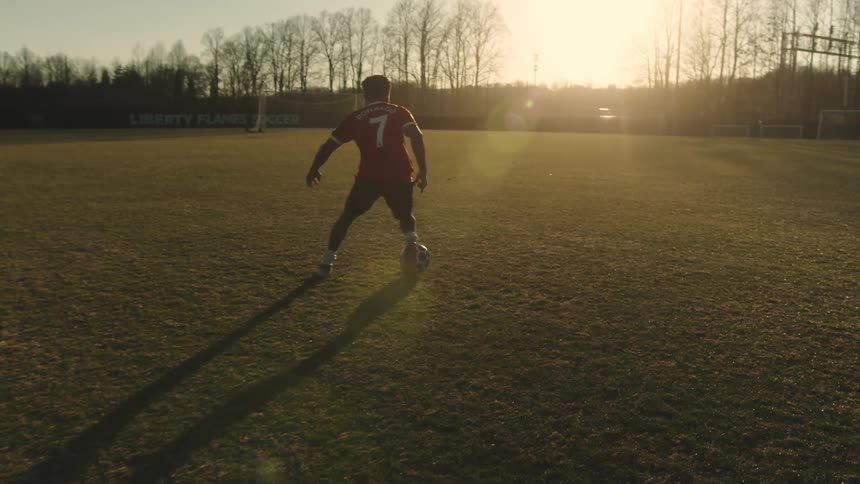 Boy dribbling soccer on a grass field with the sun on the horizon