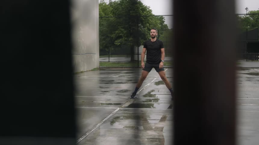 A tall man stands in a parking lot and stretches his legs on a cloudy summer day