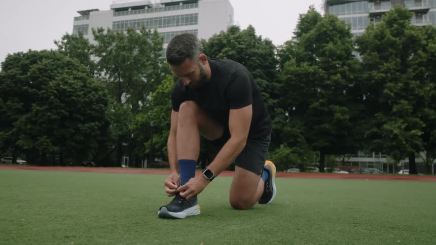 Man crouching down tying his sneakers while standing in a field with trees and buildings in the background