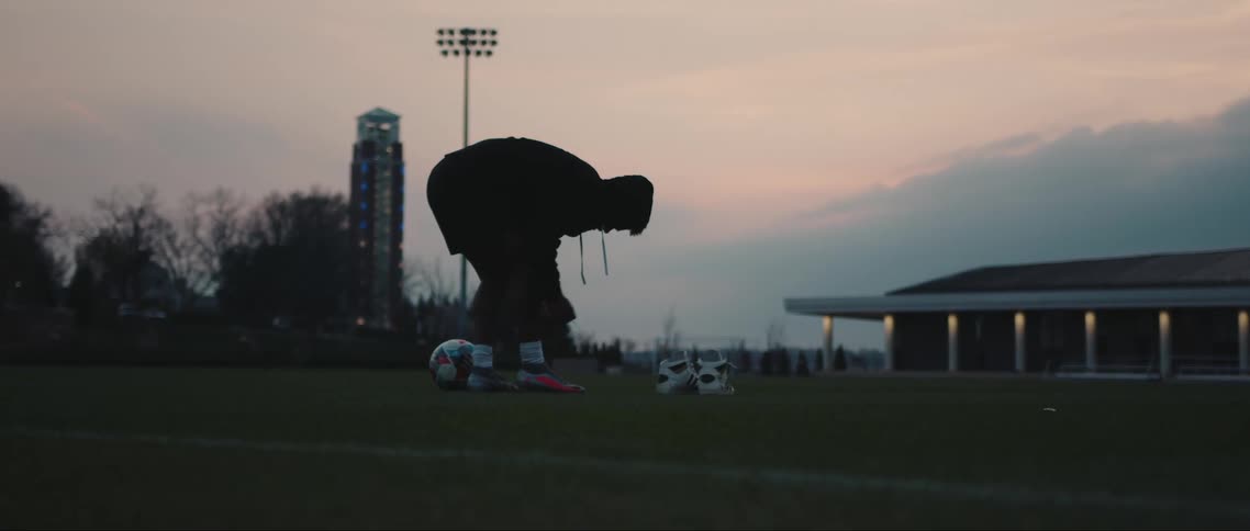 Silhouette of a boy tying his football boots on a soccer field with a ball at his side