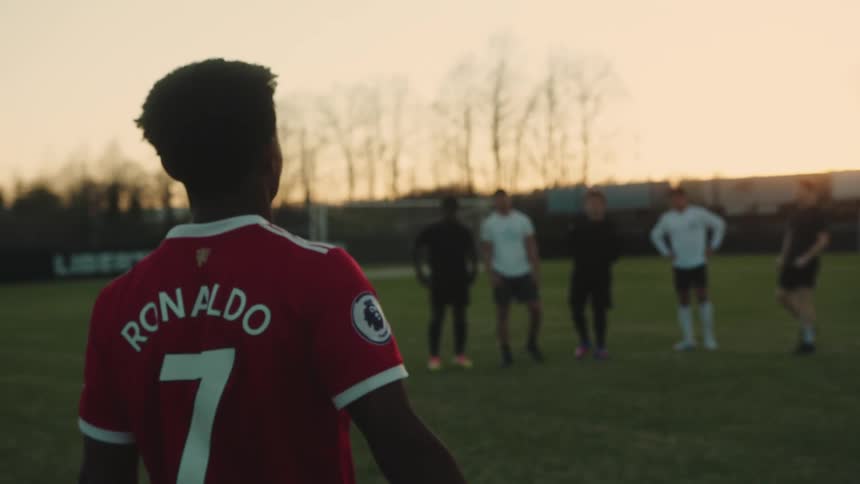 A young black player approaches five other players on a soccer field at dusk