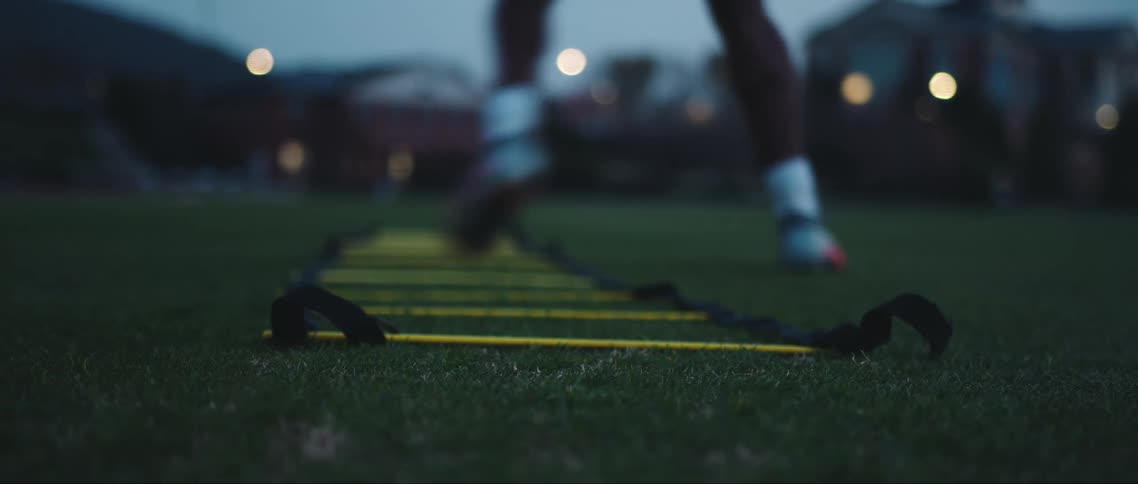 Yellow and black agility ladder in the grass with a persons foot wearing a soccer cleat passing by