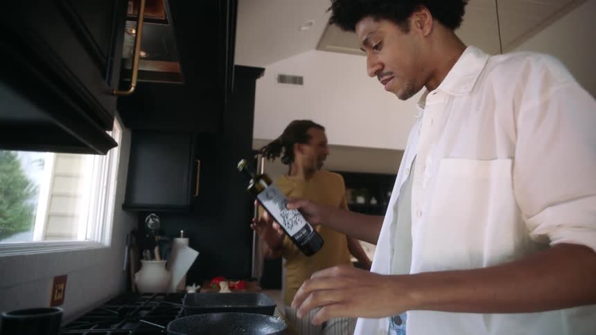 Two men in the kitchen with one of them pouring olive oil into the frying pan