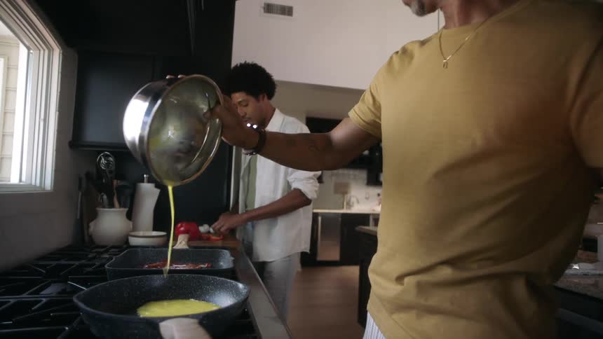 Two men in the kitchen with one of them pouring scrambled eggs into a frying pan
