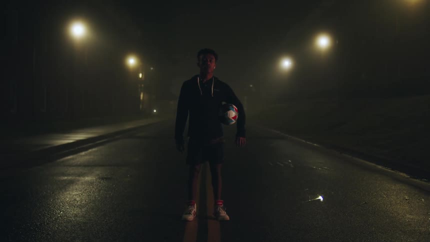A man holding a soccer ball is walking in the middle of an empty street at night