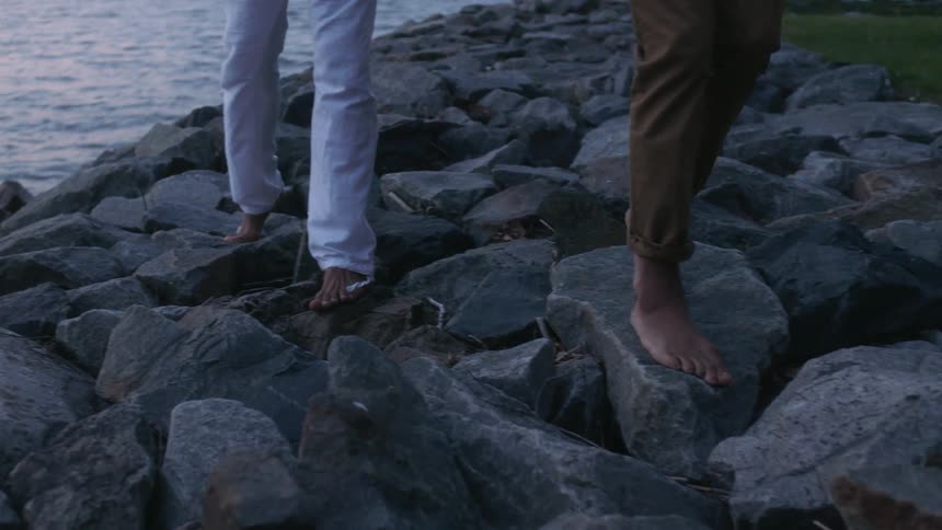 Two people in pants walking barefoot on several rocks near the sea